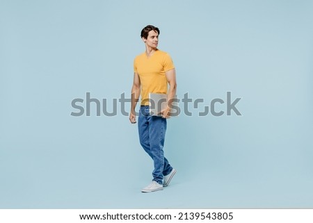 Full body happy young man 20s wear yellow t-shirt hold closed laptop pc computer look aside on workspace area mock up isolated on plain pastel light blue background studio. People lifestyle concept.