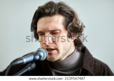 Close-up of young man singing a song to microphone with his eyes closed against the grey background