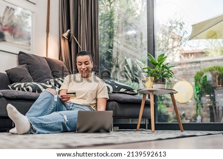 Smiling adult short haired woman, sitting on floor, looking at her bank account online.