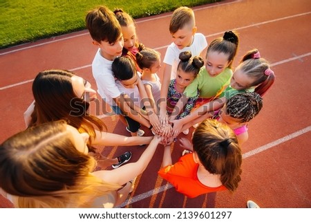 A large group of children, boys and girls, stand together in a circle and fold their hands, tuning up and raising team spirit before the game at the stadium during sunset. A healthy lifestyle. Royalty-Free Stock Photo #2139601297