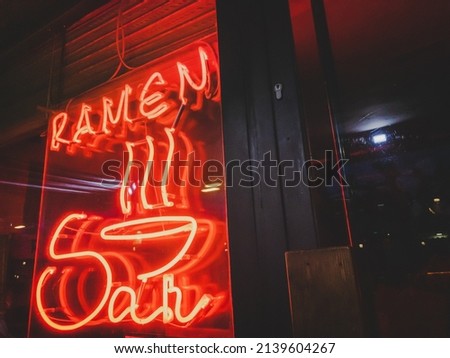 Red neon lights that have the word ramen written next to a door of a city establishment. Letters on a sign indicating that ramen soup is for sale next to a closed business.