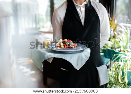 Close-up of a young waiter in a stylish uniform carrying an exquisite salad to a client in a beautiful gourmet restaurant. Table service in the restaurant. Royalty-Free Stock Photo #2139608903