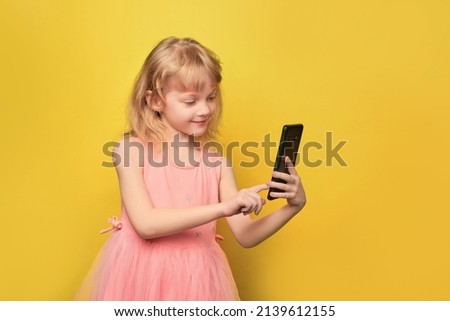 Close-up portrait of her. She is a sweet, attractive preschool girl. Smiling child holding a smartphone and taking a selfie. Curly blonde 5 years old in a pink dress on a bright yellow background.