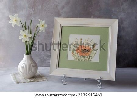 Embroidered poppy flower in a wooden white frame with a green passe-partout next to the table is a vase with daffodils