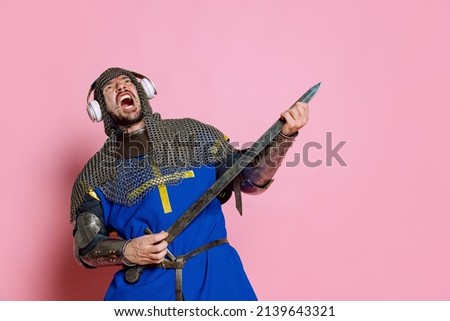 Portrait of man, medieval warrior or knight in armor with headphones holding sword like guitar and playing isolated over pink background. Rock performer. Comparison of eras, history, renaissance style