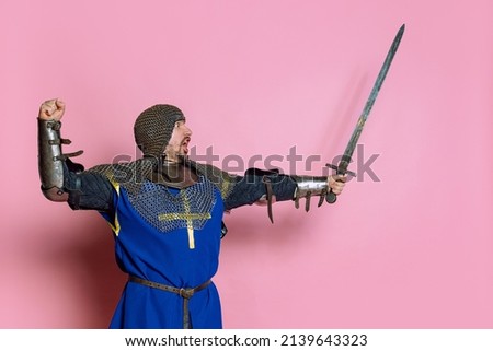 Portrait of brave man, medieval warrior or knight in armor rising sword, showing readiness to fight isolated over pink studio background. Comparison of eras, history, renaissance style
