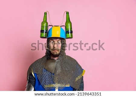 Portrait of funny looking man, medieval warrior or knight in beer helmet and protective armor isolated over pink studio background. Beer lover. Comparison of eras, history, renaissance style, festival