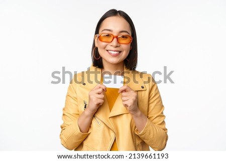 Portrait of smiling korean female model in sunglasses, showing credit card, standing over white background