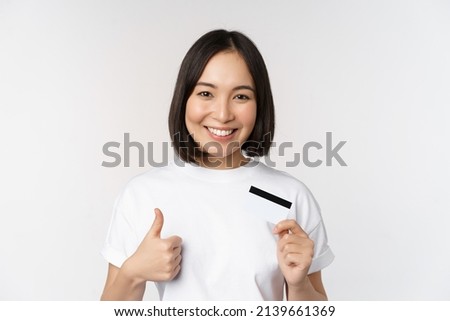 Portrait of beautiful young modern asian woman, showing credit card and thumbs up, recommending contactless payment, standing over white background