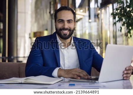 Handsome smiling Moroccan businessman using laptop computer working online sitting in modern office. Portrait of happy successful middle eastern manager looking at camera sitting at workplace 