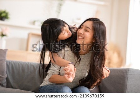 Fun mom with cute baby girl. Mother and daughter playing together in the living room. Asian girl embracing mom from stand behind while mom sitting on couch. Happy family, motherhood, childhood concept