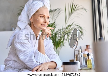 Beautiful mid age adult 50 years old blonde woman wears bathrobe and towel in bathroom applying rejuvenating tightening antiage face skin care cream treatment, sitting at mirror. Daily beauty routine. Royalty-Free Stock Photo #2139673569