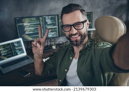Photo of funny cheerful young man wear green shirt spectacles recording video showing v-sign indoors workshop workplace.