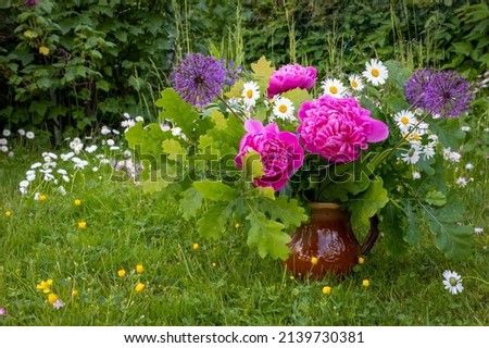 A bouquet of pink peonies, daisies, decorative onion flowers and oak twigs in a brown clay vase on a natural green background. Midsummer bouquet.