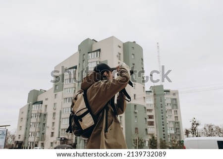 Portrait of a young male photographer with a camera taking photos or videos on the street in the yard. Handsome young man with a professional camera on the background of an apartment building