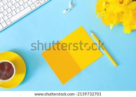 Woman's modern workspace. Keyboard, and office supplies on blue table. Bouquet of delicate beautiful yellow daffodils and cup of tea for holiday. 
Flat lay  top view  concept with copy space for text.
