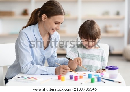 Art development. Cute little boy and his young mother drawing together with colorful paints, sitting at classroom and smiling