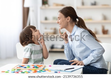 Speech training for kids. Professional woman specialist training with little boy at cabinet, teaching him right articulation exercises, side view Royalty-Free Stock Photo #2139736355