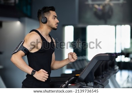 Muscular Arab Male Athlete Wearing Wireless Headphones Running On Treadmill At Gym, Sporty Millennial Middle Eastern Guy Listening Music During Workout, Enjoying Cardio Training, Copy Space Royalty-Free Stock Photo #2139736545