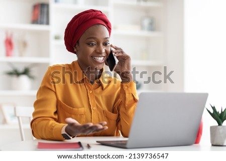 Positive young muslim black lady with red turban on her head and casual outfit working on laptop at office, looking at computer screen and talking on cell phone with business partner, copy space Royalty-Free Stock Photo #2139736547
