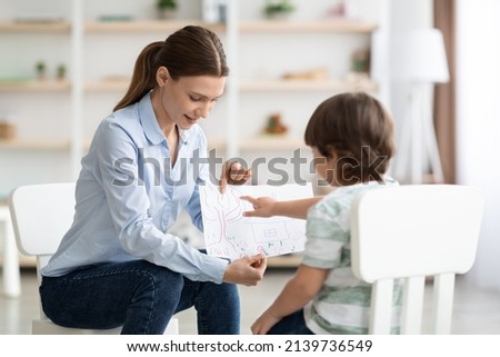Kids psychology concept. Professional woman psychologist examining little patient at office, boy describing picture with nature during consultation, free space