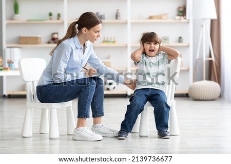 Difficult kid concept. Emotional angry little boy screaming and covering ears during consultation at psychologist, worried therapist trying to calm down patient at office Royalty-Free Stock Photo #2139736677