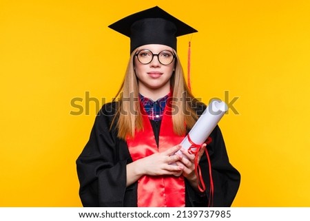 Girl graduate in graduation hat and eyewear with diploma on yellow background. Blonde young woman wearing graduation cap and ceremony robe holding Certificate tied with red ribbon. Education Concept Royalty-Free Stock Photo #2139737985