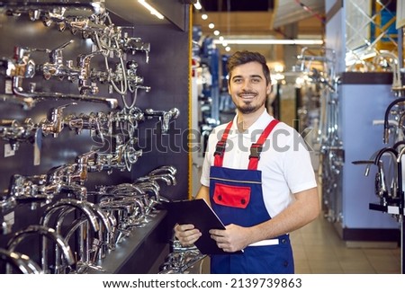 Portrait of a young man who sells faucets. Happy salesman in uniform standing in the aisle with modern water taps at a big hardware store or shopping mall Royalty-Free Stock Photo #2139739863