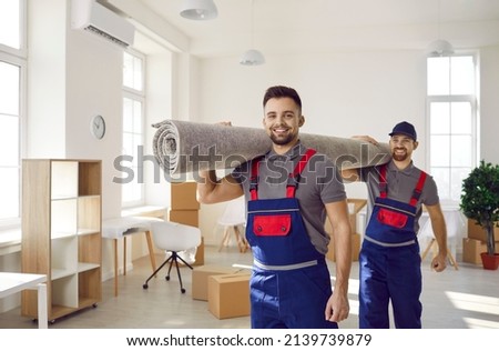 Smiling young male movers carrying carpet unload goods in client home or office. Happy removal company male workers unpack help client with boxes during moving or relocation. Transportation. Royalty-Free Stock Photo #2139739879