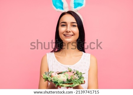Happy woman in rabbit ears holding a green wreath with colored eggs, a traditional holiday, in the studio on a pink background, Easter mood. high quality photo