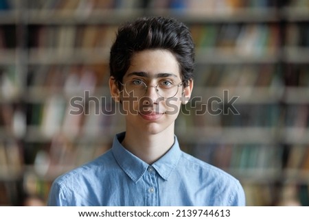 Head shot schoolboy guy posing in campus library on bookshelves background. 17s pupil in eyeglasses look at cam. Excellent student portrait, skill and knowledge, education, generation Z person concept