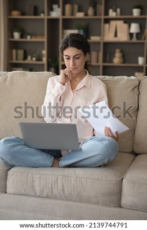 Serious thoughtful freelancer woman doing business paperwork at home, working laptop computer on couch. Renter girl paying domestic bills, taxes, using online app for online payment. Vertical shot Royalty-Free Stock Photo #2139744791