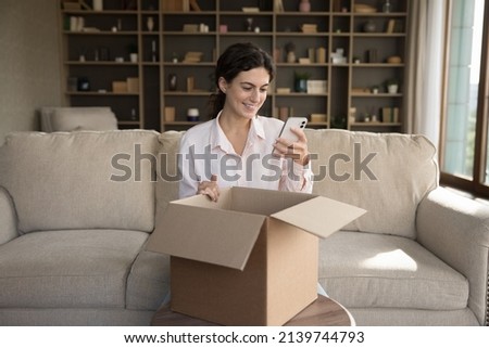 Happy positive online store customer giving feedback to purchase, internet shop, using app on mobile phone, ordering parcel delivery for sending. Young client woman unpacking carton box on sofa Royalty-Free Stock Photo #2139744793