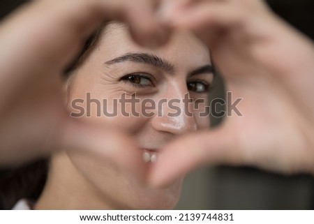 Happy beautiful millennial Latin girl looking at camera through hand heart shaped frame, joining fingers, smiling. Cheerful blogger woman expressing love, support, gratitude, showing like. Close up Royalty-Free Stock Photo #2139744831