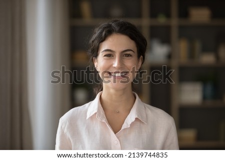 Confident happy beautiful Hispanic student girl indoor head shot portrait. Brunette young woman wearing pale natural linen shirt, smiling at camera, showing perfect teeth. Front view