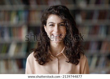 Close up attractive brunette teenage girl with curly hair pose in library look at camera, head shot portrait on bookshelves background. Higher education institution student, excellent studies concept