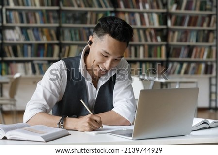 Handsome Asian guy hold pencil take notes in copybook while studying alone in library. Student wear earbuds listen audio task, makes assignment sit at table. Education, improve knowledge, tech concept Royalty-Free Stock Photo #2139744929