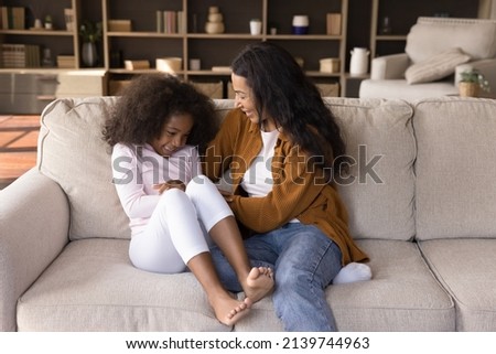Joyful happy African mom tickling excited daughter girl on couch, playing with child at home, enjoying active games, motherhood, having fun, laughing, giggling. Family, parenthood concept.