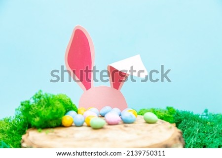 The pink rabbit hides behind a stump. Easter greeting card with Easter symbols.
