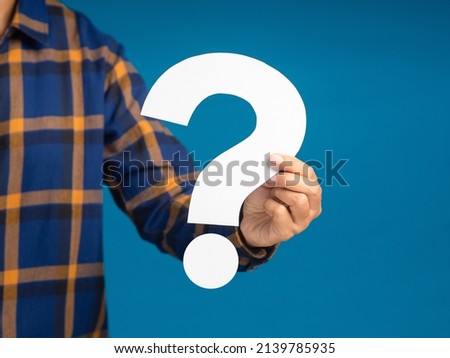Question mark symbol concept. Hand holding a white question mark paper while standing on a blue background in the studio. Close-up photo. Space for text