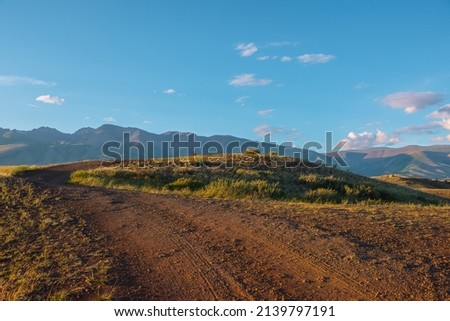 Tourist on sunlit gold grassy hill with dirt road with view to high mountain range. Beautiful sunny mountain landscape with man in mountains. Colorful scenery with traveler on hill in golden sunlight.