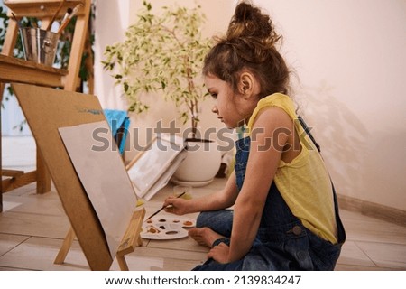 Adorable European child, cute little girl sitting on the floor at a wooden easel and drawing picture painting on canvas. Art class, creativity, children entertainment concept.