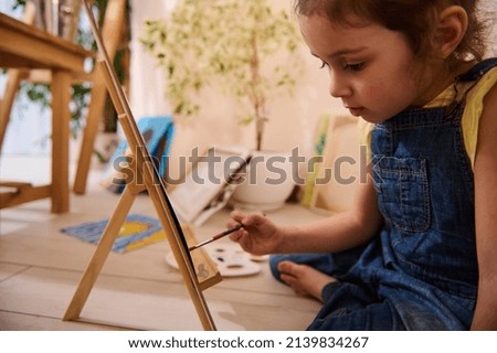 Adorable Caucasian child, cute little girl sitting on the floor at a wooden easel and drawing picture painting on canvas. Art class, creativity, children entertainment concept.