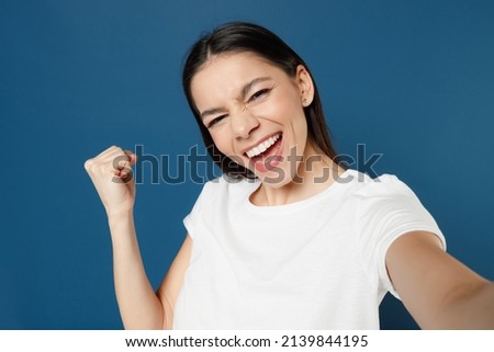 Close up young overjoyed excited happy latin woman 20s in white casual basic t-shirt doing selfie shot on mobile phone show winner gesture clench fist isolated on dark blue background studio portrait