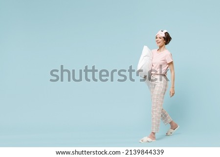 Full length young smiling caucasian woman in pajamas jam sleep eye mask rest relax at home holding pillow walking going isolated on pastel blue color background studio. Good mood night bedtime concept Royalty-Free Stock Photo #2139844339