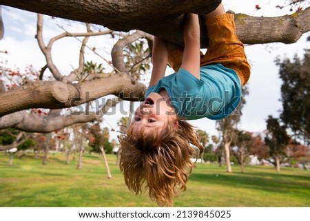 Cute blonde child boy hangs on a tree branch. Summer holidays, little boy climbing a tree. Upside down. Children love nature on countryside. Royalty-Free Stock Photo #2139845025