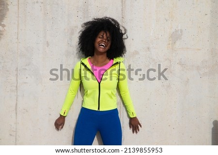 Beautiful young Afro American woman in bright green and pink sportswear on a grey concrete wall texture background. Woman makes different expressions. Laughing, serious, happy, sad, thinking