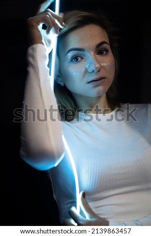 The portrait of beautiful woman in white clothes, top, shiny shimmer makeup and stars with blue led stripes, neon light on black background. Futuristic style girl, sadly emotions. Future, amazing.