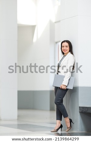 Portrait of successful Asian businesswoman in a relaxed business suit holds laptop computer between business buildings
