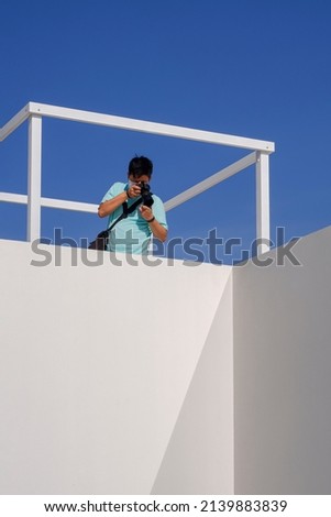 Young Asian man taking picture with digital camera on rooftop of white building against blue clear sky in vertical frame